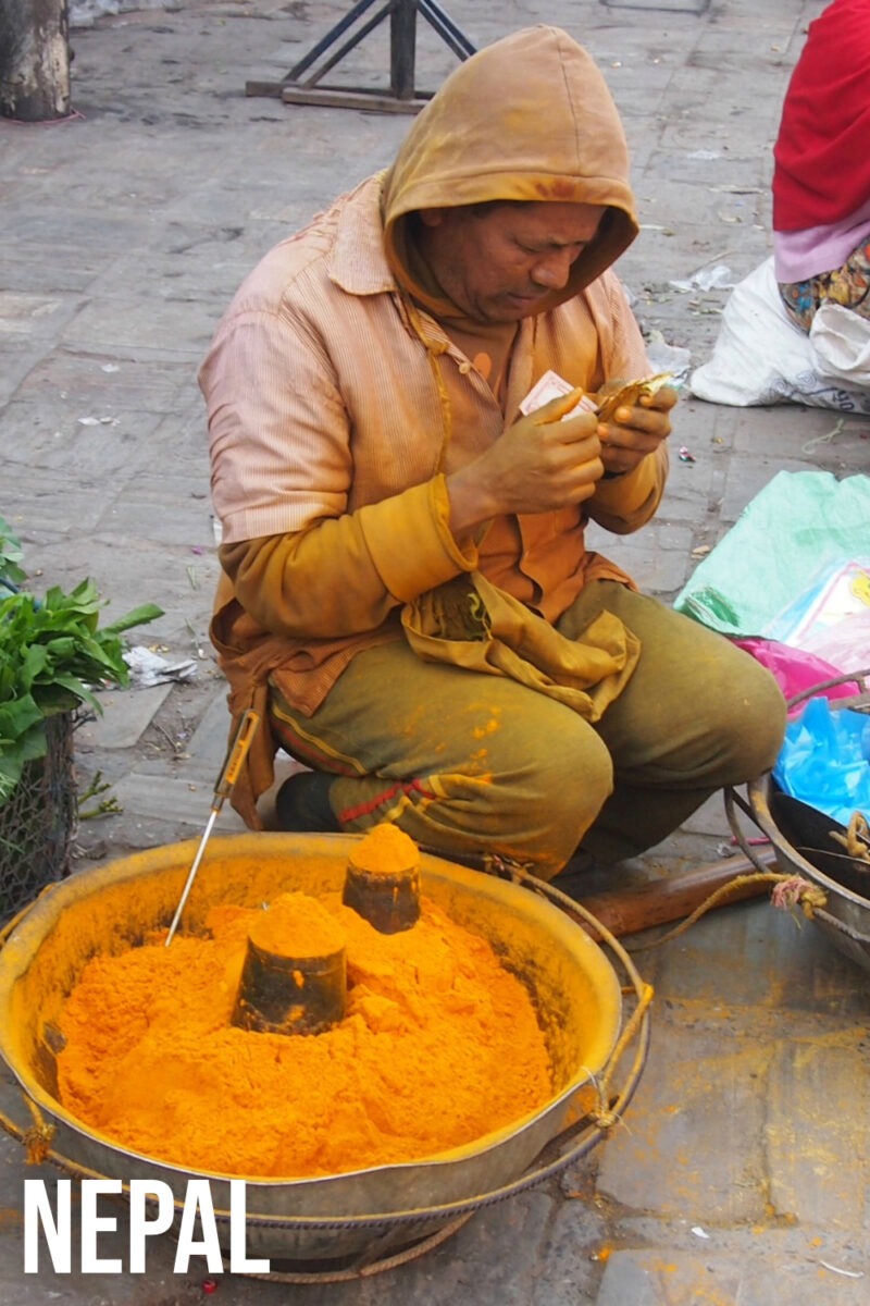 A man selling turmeric, stained in yellow, from a basket at a street market in Nepal