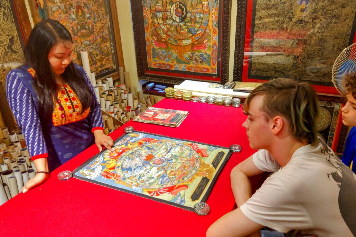 Thanka shop. Kids look at a thanka while a woman explains the religious significance. A thanka is on the table and many are on the walls.